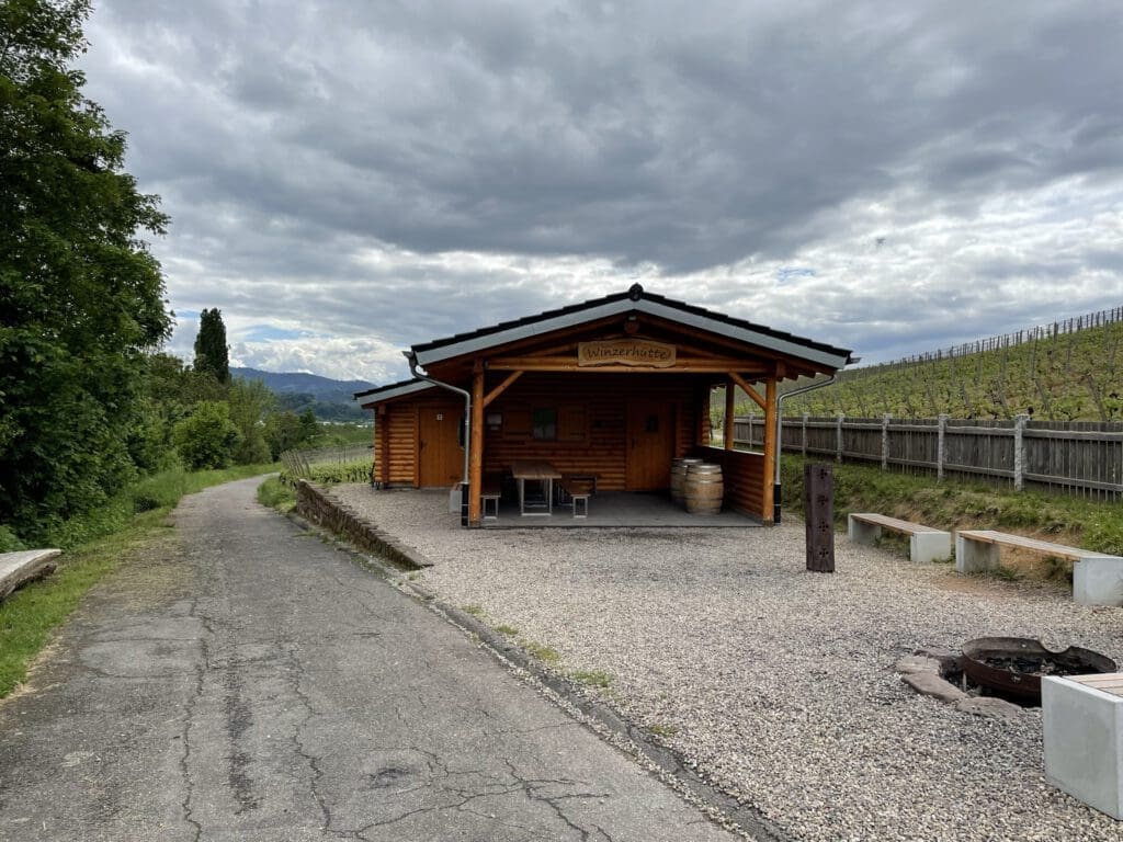 Eine kleine Holzhütte, die einer traditionellen Grillhütte mit überdachter Veranda ähnelt, steht an einem Kiesweg. Im Vordergrund stehen zwei Bänke und eine leere Feuerstelle. Rechts erstreckt sich unter dem wolkenverhangenen Himmel ein Weinberg entlang eines Hügels. Das Bild dient auch als Referenz für Trockentrenntoiletten in Hütten.