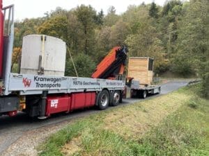 Auf dem Foto ist ein LKW mit einer Goldgrube drauf. Dieser hat auch einen Anhänger mit einem Toilettenhaus aus Holz drauf. Der Laster steht auf einer kleinen Straße mit Wiesen und Wäldern im außen herum