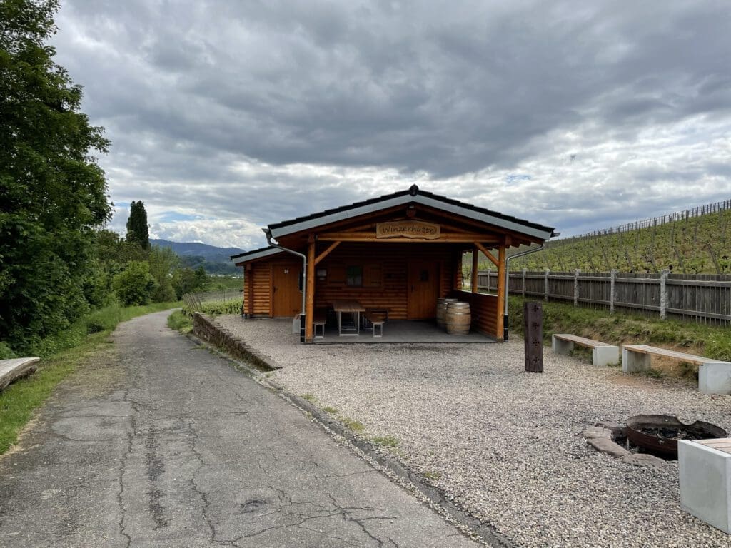 Eine kleine Holzhütte, die einer traditionellen Grillhütte mit überdachter Veranda ähnelt, steht an einem Kiesweg. Im Vordergrund stehen zwei Bänke und eine leere Feuerstelle. Rechts erstreckt sich unter dem wolkenverhangenen Himmel ein Weinberg entlang eines Hügels. Das Bild dient auch als Referenz für Trockentrenntoiletten in Hütten.