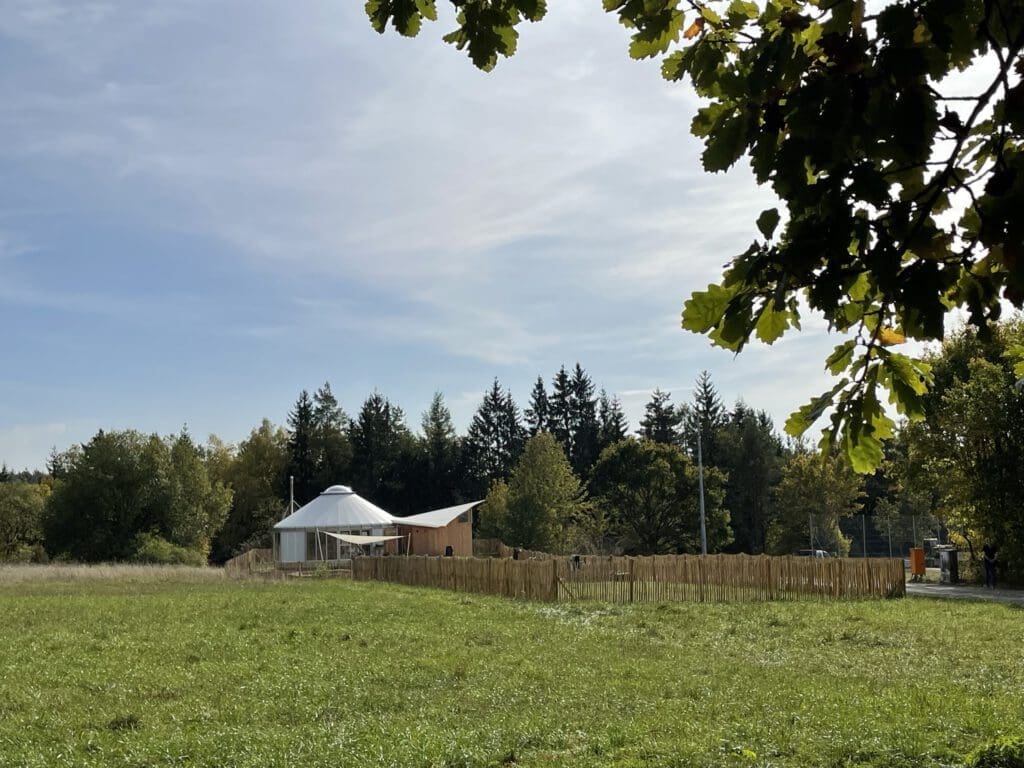 Eine Jurte mit einem Holzzaun steht auf einer Wiese unter blauem Himmel und erinnert an einen Waldkindergarten. Bäume säumen das Gelände, und große Blätter rahmen die Szene von oben ein und schaffen eine friedliche und natürliche Landschaft. Das Bild dient auch als Referenz für Trockentrenntoiletten bei Kindergärten.