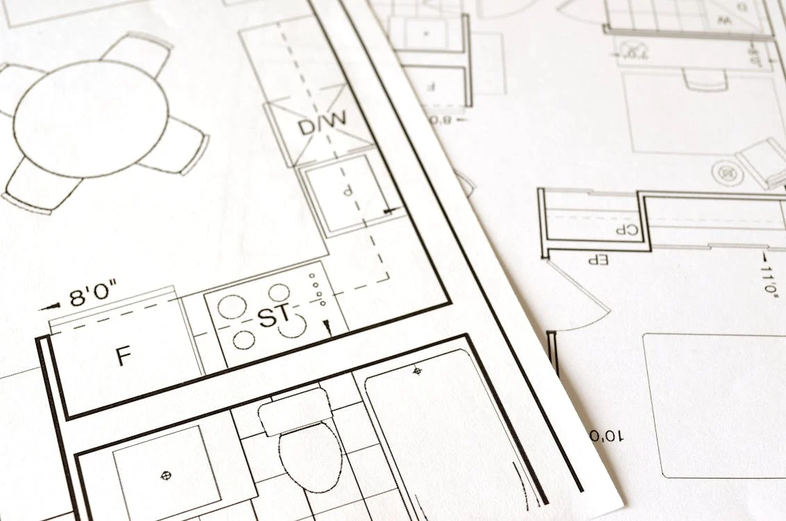 Eine Nahaufnahme architektonischer Pläne, die einem komplizierten Datenblatt ähneln, zeigt einen Grundriss einer Küche mit Herd und Spüle sowie eines Badezimmers mit Toilette. Die Pläne sind elegant über eine makellos weiße Oberfläche verteilt. Das Bild soll unsere Sammlung an Datenblätter n darstellen.