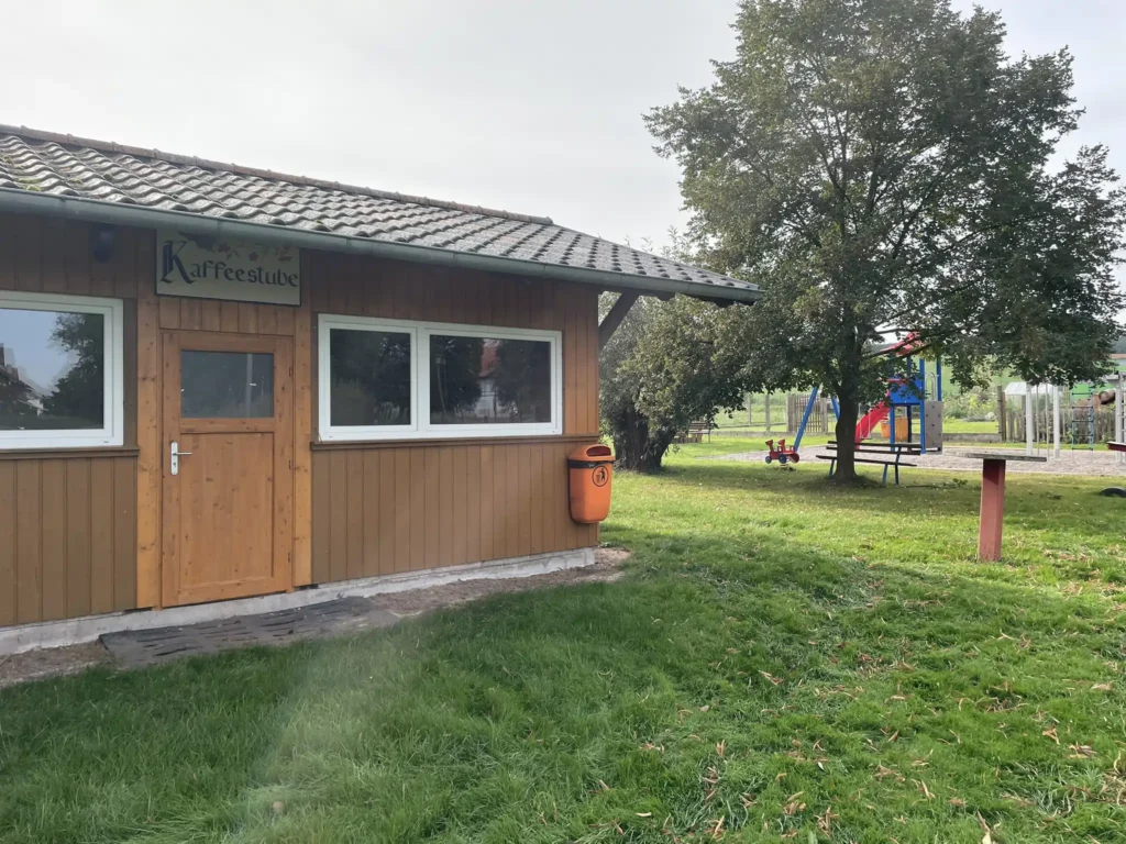 Auf dem Bild ist eine "Kaffeestube" im Vordergrund, während ein Spielplatz und ein Baum im Hintergrund zu sehen ist. Die Sonne scheint auf das Objektiv und das Grass ist saftig grün. Hier wurde ein LEADER Projekt umgesetzt.