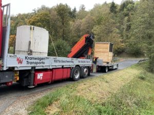 Auf dem Foto ist ein LKW mit einer Goldgrube drauf. Dieser hat auch einen Anhänger mit einem Toilettenhaus aus Holz drauf. Der Laster steht auf einer kleinen Straße mit Wiesen und Wäldern im außen herum