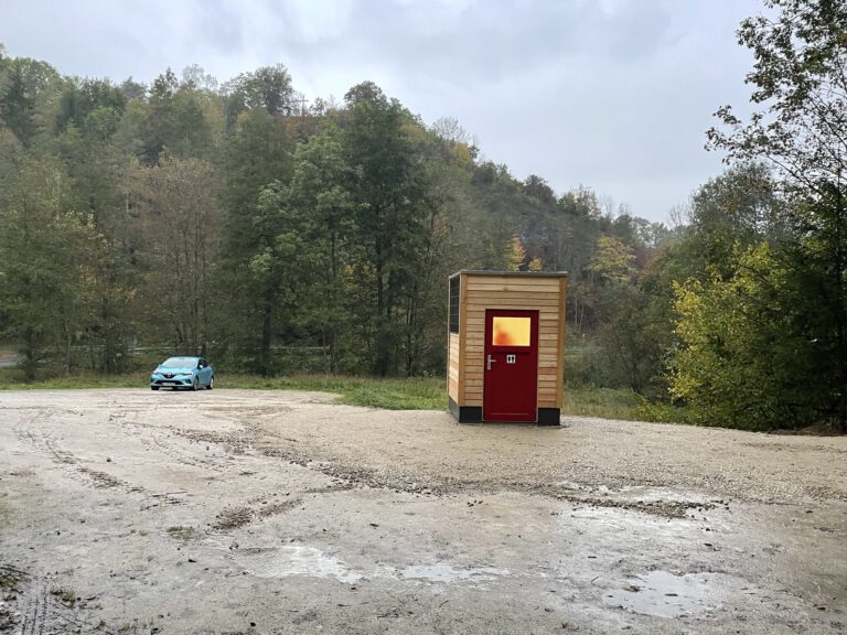 Auf dem Foto ist ein Toilettenhaus LÄRCHE im Ahorntal. Dahinter sieht man Bäume und davor den Parkplatz. Das Toilettenhaus hat eine rote Türe und das Licht ist an.